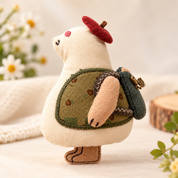 Handcrafted cream-colored cartoon animal plush brooch with a red beret, olive-green outfit, and miniature backpack