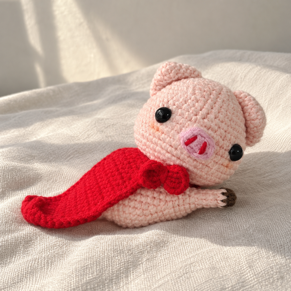 Pink crochet pig plush reclining on cream fabric with a bright red cape and matching bow