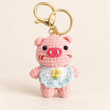 Pink crochet pig keychain wearing a pale blue outfit with a yellow daisy and gold-tone clasp